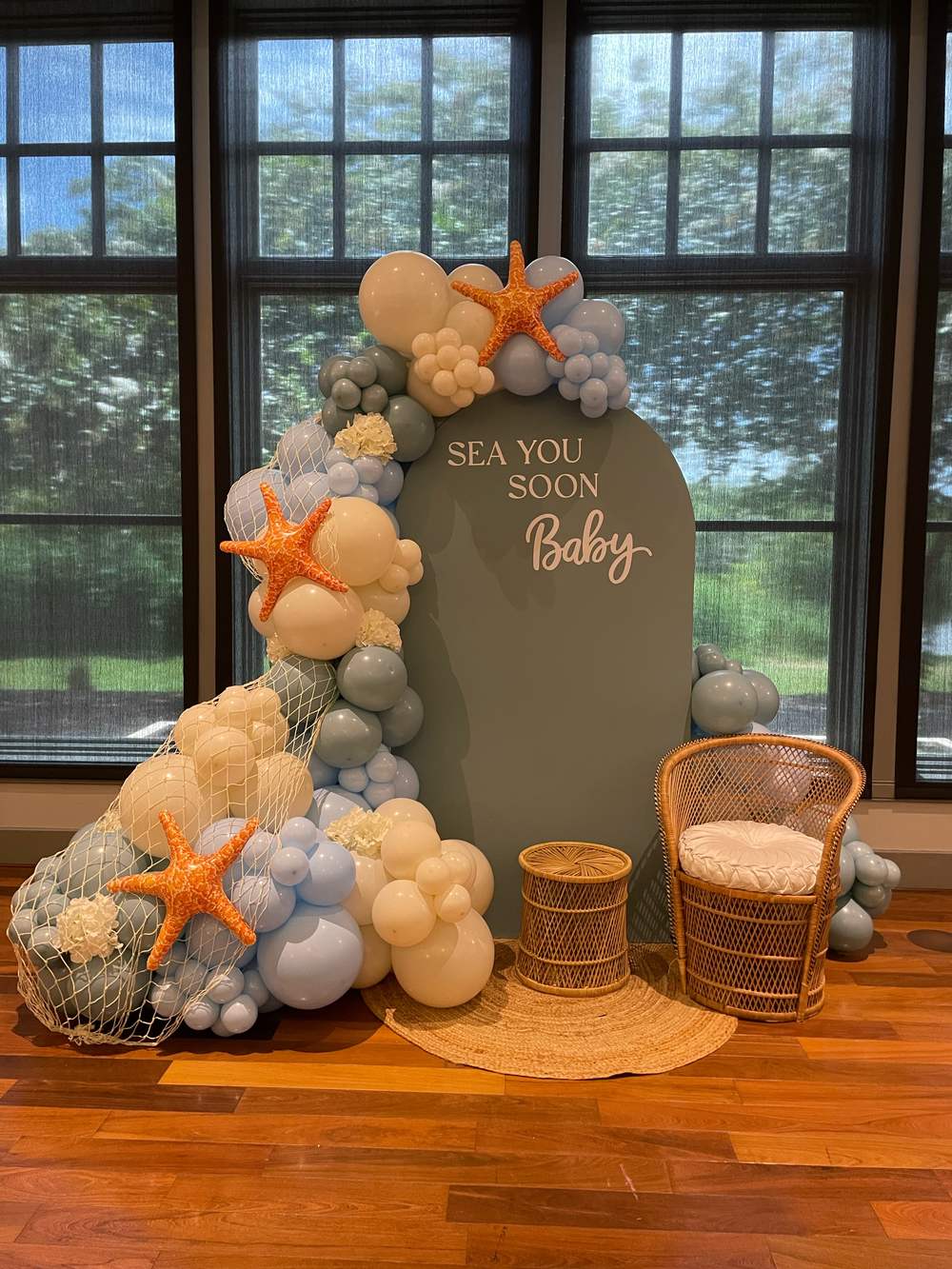 The dressed arch panel with the balloon garland beside it, a rattan peacock chair and a small rattan side table set on a round jute rug.