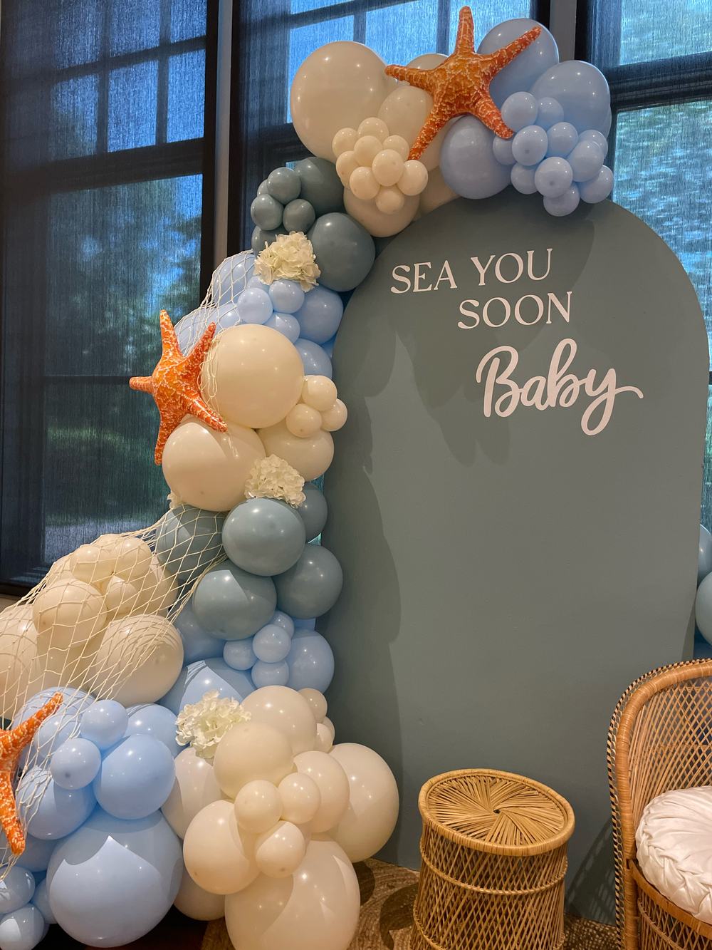 Close view of the garland: cream, sand, dusty blue and sage balloons in mixed sizes, with fishnet draped across them, orange starfish and dried flowers tucked into the cluster.