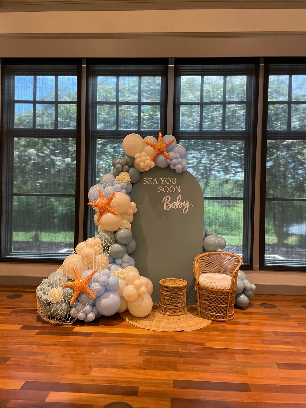 The full installation seen from across the room: a sage arch panel against a wall of tall windows, a cascading balloon garland in cream, sand and dusty blue down its left side, with rattan seating and a jute rug in front.